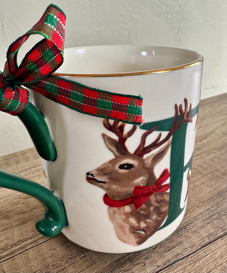 Puede incluir: Taza de cer&aacute;mica blanca con asa verde y borde dorado. La taza presenta una ilustraci&oacute;n de ciervo con un lazo rojo y la letra "E". Una cinta a cuadros roja, verde y dorada est&aacute; atada alrededor del asa.