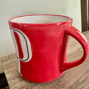 May include: A red ceramic mug with a white "P" design. The mug has a curved handle and a white interior. The mug is sitting on a wooden surface.
