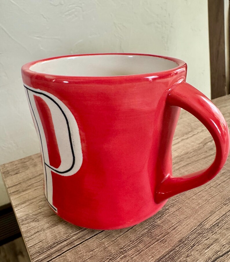 May include: A red ceramic mug with a white "P" design. The mug has a curved handle and a white interior. The mug is sitting on a wooden surface.