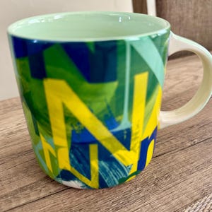 May include: A ceramic mug with a green interior and handle. The exterior features a vibrant abstract design in shades of green, blue, and yellow. The mug is set on a wooden surface.