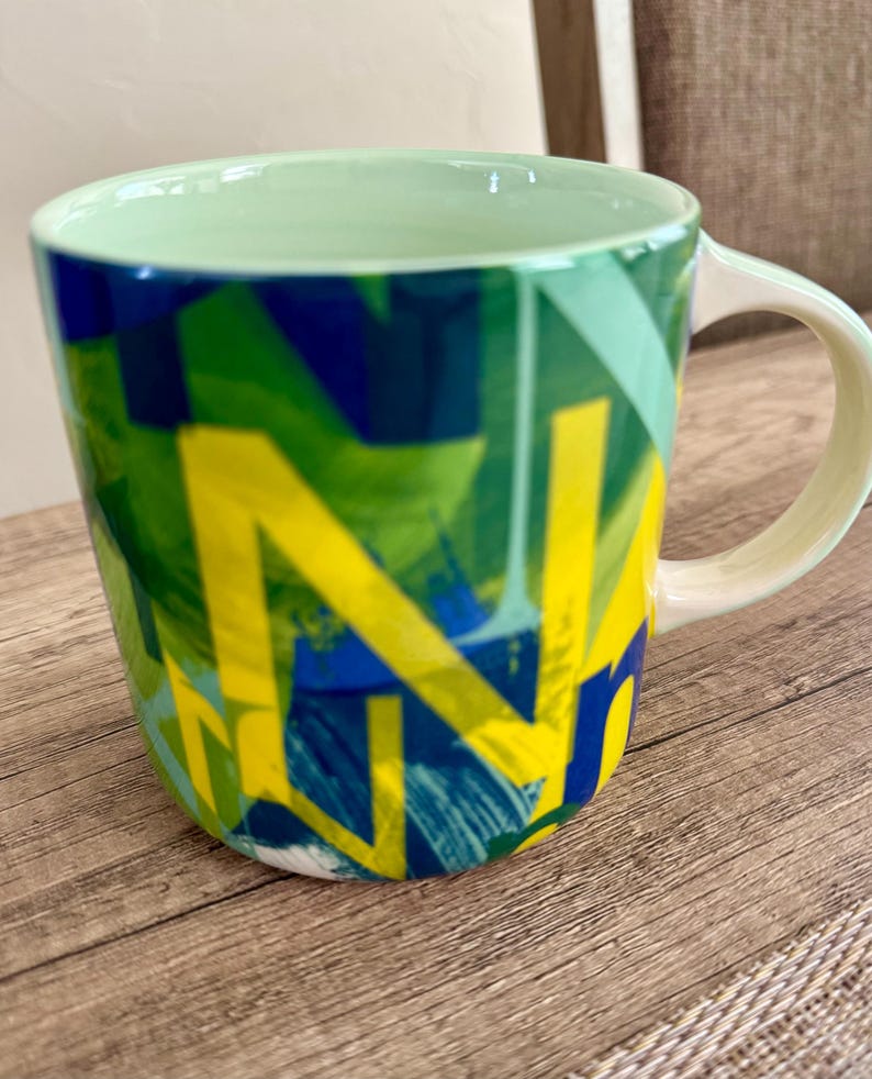 May include: A ceramic mug with a green interior and handle. The exterior features a vibrant abstract design in shades of green, blue, and yellow. The mug is set on a wooden surface.