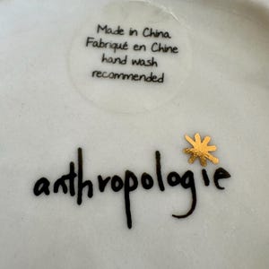 May include: A white ceramic surface featuring the black text Made in China, Fabriqu&eacute; en Chine, and hand wash recommended. Below this, the word anthropologie is printed in black script with a small, metallic gold star-shaped icon above the letter i.