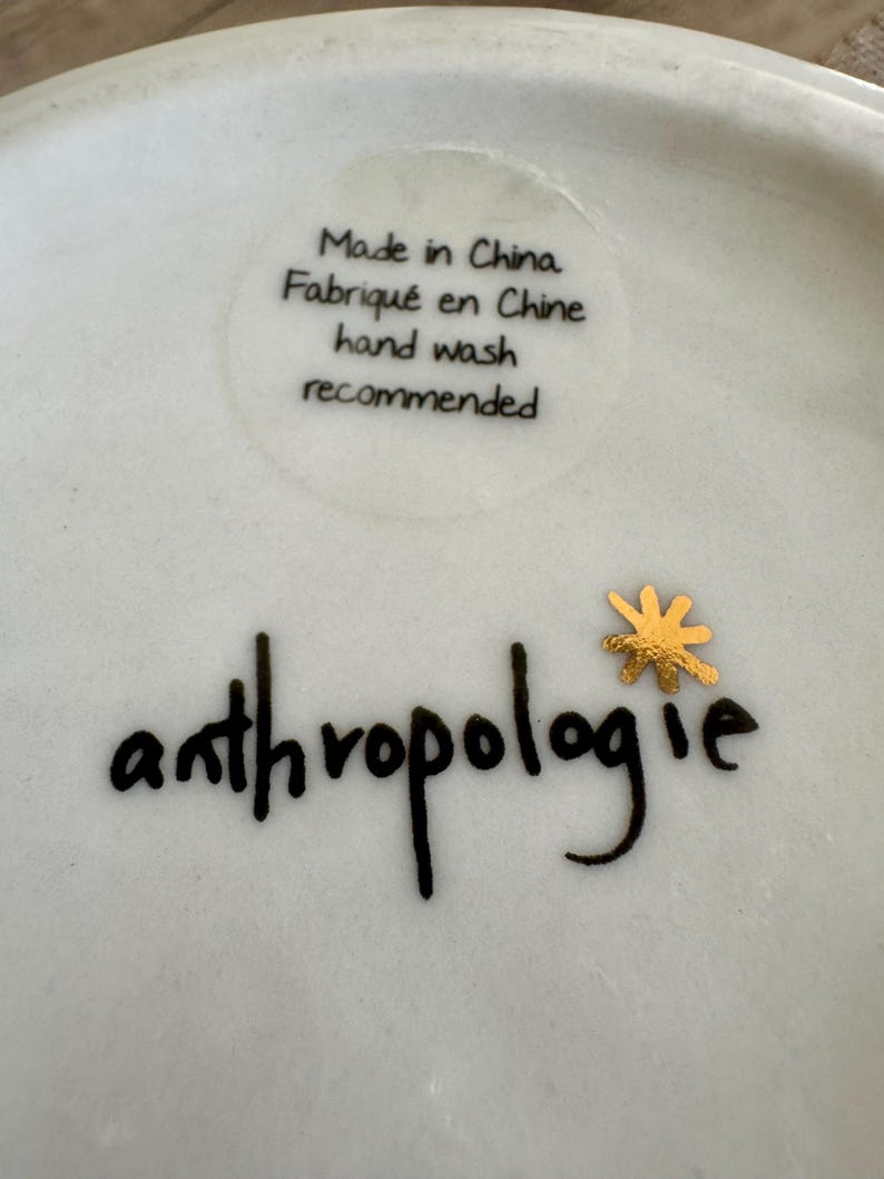 May include: A white ceramic surface featuring the black text Made in China, Fabriqu&eacute; en Chine, and hand wash recommended. Below this, the word anthropologie is printed in black script with a small, metallic gold star-shaped icon above the letter i.