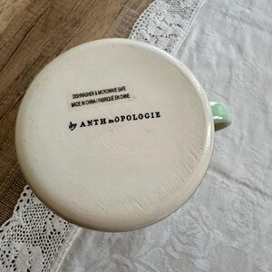 May include: Off-white ceramic mug with a light green handle. The bottom of the mug features the text "DISHWASHER & MICROWAVE SAFE MADE IN CHINA / FABRIQUE EN CHINE" and "by ANTHROPOLOGIE".