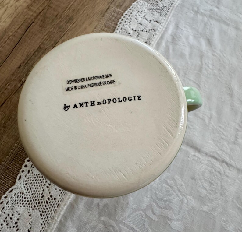 May include: Off-white ceramic mug with a light green handle. The bottom of the mug features the text "DISHWASHER & MICROWAVE SAFE MADE IN CHINA / FABRIQUE EN CHINE" and "by ANTHROPOLOGIE".