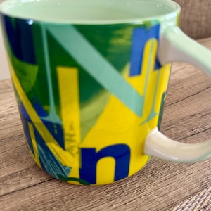 May include: A ceramic coffee mug with a green interior and handle. The exterior features a vibrant design with yellow, blue, and green geometric shapes and letters. The mug is set on a wooden surface.