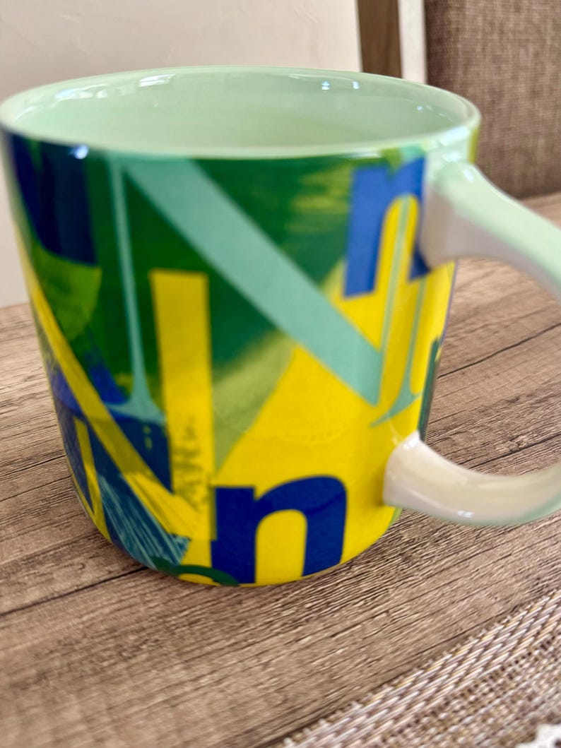 May include: A ceramic coffee mug with a green interior and handle. The exterior features a vibrant design with yellow, blue, and green geometric shapes and letters. The mug is set on a wooden surface.