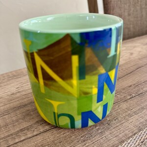 May include: A ceramic mug with a light green interior and a colorful exterior design. The exterior features a mix of green, yellow, and blue geometric shapes with the letter "N" in yellow and blue.