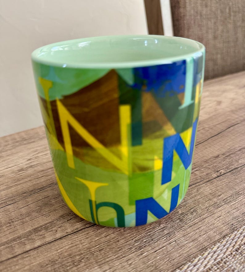 May include: A ceramic mug with a light green interior and a colorful exterior design. The exterior features a mix of green, yellow, and blue geometric shapes with the letter "N" in yellow and blue.
