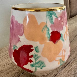 May include: A white ceramic vase with a gold rim, decorated with colourful floral patterns in red, pink, and orange. The vase has a slightly tapered shape and is set against a wooden surface.