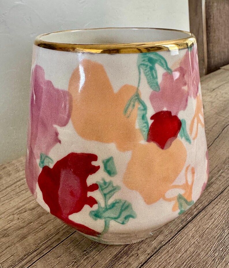 May include: A white ceramic vase with a gold rim, decorated with colourful floral patterns in red, pink, and orange. The vase has a slightly tapered shape and is set against a wooden surface.