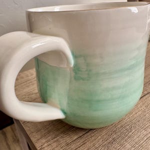Pode incluir: Uma caneca de cer&acirc;mica com a parte superior branca e um design verde em estilo aquarela na parte inferior. A caneca tem uma al&ccedil;a grande e curva e est&aacute; sobre uma superf&iacute;cie de madeira. A caneca &eacute; de cor pastel clara.