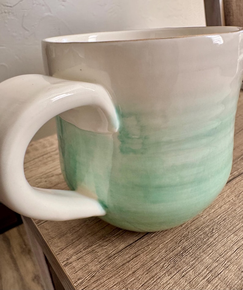 Pode incluir: Uma caneca de cer&acirc;mica com a parte superior branca e um design verde em estilo aquarela na parte inferior. A caneca tem uma al&ccedil;a grande e curva e est&aacute; sobre uma superf&iacute;cie de madeira. A caneca &eacute; de cor pastel clara.