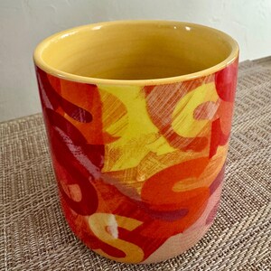 May include: A cylindrical ceramic container with a yellow interior and rim. The exterior features a vibrant abstract design in shades of red, orange, and yellow. The container is likely used for holding items.