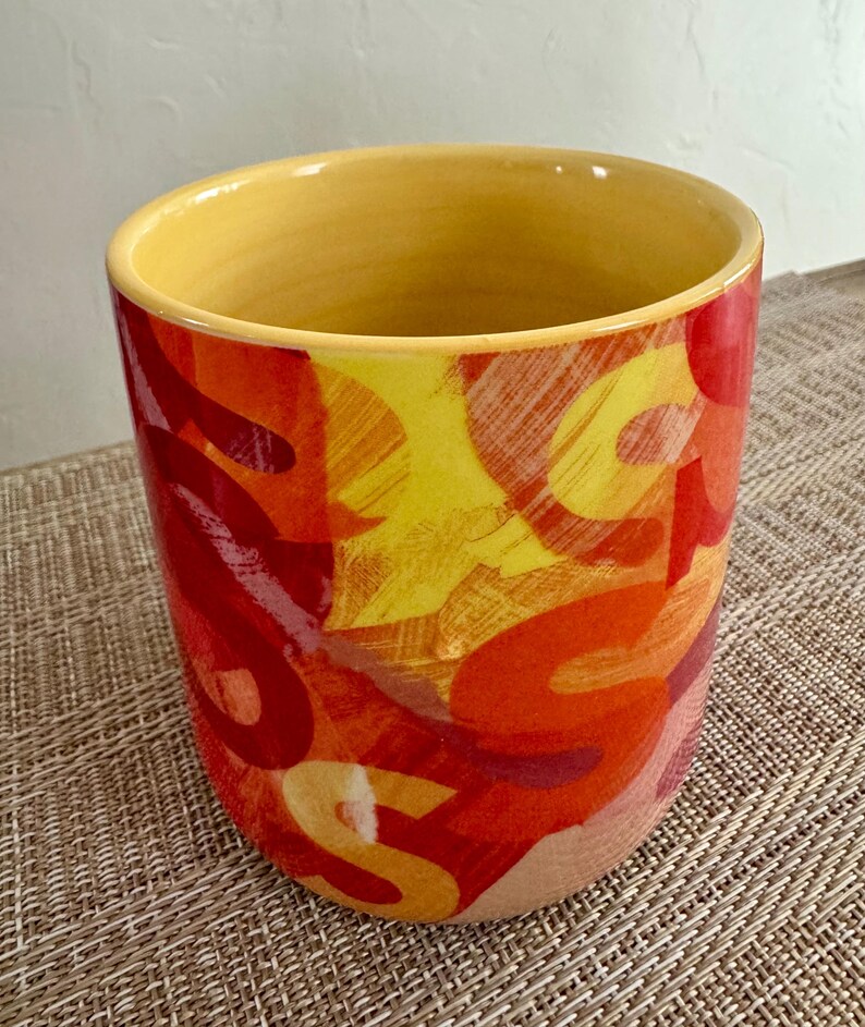 May include: A cylindrical ceramic container with a yellow interior and rim. The exterior features a vibrant abstract design in shades of red, orange, and yellow. The container is likely used for holding items.