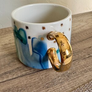 May include: A white ceramic mug featuring a blue and teal watercolor-style abstract pattern and small gold star accents. The mug has a large, reflective gold-toned handle that curves outward from the side, providing a distinct metallic contrast.