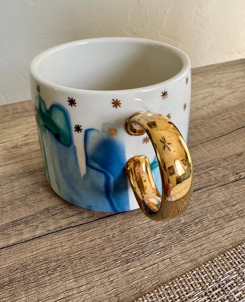 May include: A white ceramic mug featuring a blue and teal watercolor-style abstract pattern and small gold star accents. The mug has a large, reflective gold-toned handle that curves outward from the side, providing a distinct metallic contrast.