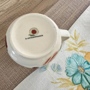 May include: White ceramic mug with a handle, decorated with red and brown floral patterns. The base of the mug has the text "by ANTHROPOLOGIE". The mug is on a floral patterned cloth.