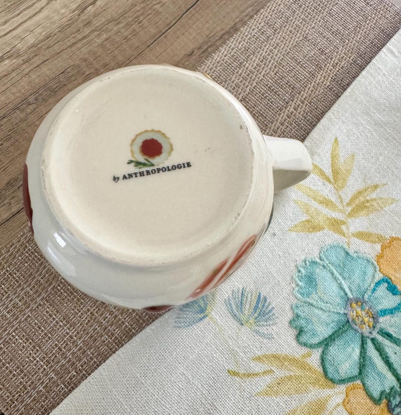May include: White ceramic mug with a handle, decorated with red and brown floral patterns. The base of the mug has the text "by ANTHROPOLOGIE". The mug is on a floral patterned cloth.