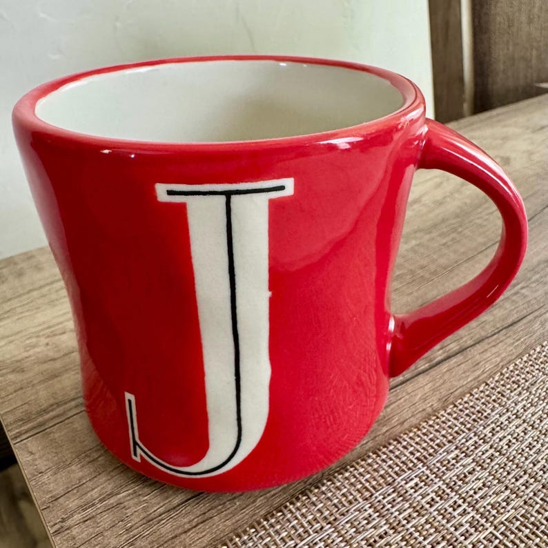 May include: A red ceramic mug with a white interior and a large white letter "J" outlined in black. The mug has a curved handle and sits on a wooden surface.