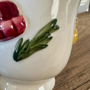 K&ouml;nnte beinhalten: Ein wei&szlig;er Keramikkrug mit gl&auml;nzendem Finish. Der Krug hat ein gr&uuml;nes Blattdesign und ein rot-wei&szlig;es Dekorationselement. Der Griff hat ein gelb-rosa Design. Der Krug steht auf einem wei&szlig;en Sockel.