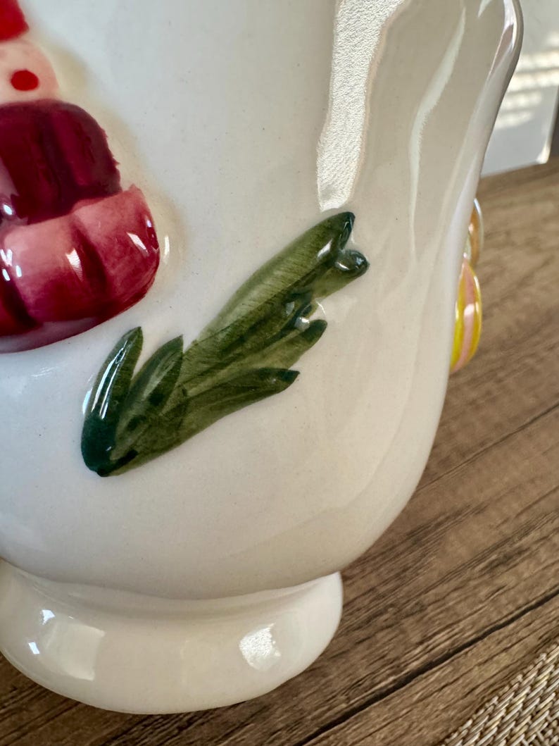 K&ouml;nnte beinhalten: Ein wei&szlig;er Keramikkrug mit gl&auml;nzendem Finish. Der Krug hat ein gr&uuml;nes Blattdesign und ein rot-wei&szlig;es Dekorationselement. Der Griff hat ein gelb-rosa Design. Der Krug steht auf einem wei&szlig;en Sockel.