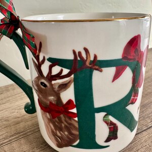 Puede incluir: Taza de cer&aacute;mica blanca con asa verde y borde dorado. La taza presenta una gran letra verde "F" con una ilustraci&oacute;n de reno marr&oacute;n y un lazo rojo. Una cinta a cuadros roja y verde est&aacute; atada al asa.