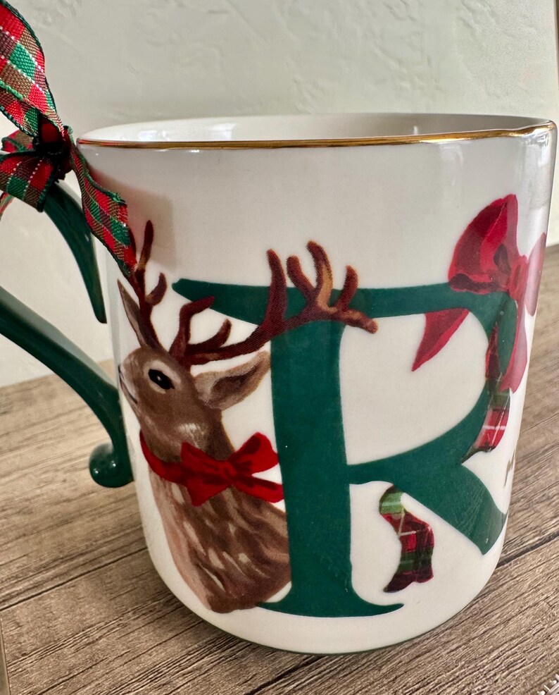 Puede incluir: Taza de cer&aacute;mica blanca con asa verde y borde dorado. La taza presenta una gran letra verde "F" con una ilustraci&oacute;n de reno marr&oacute;n y un lazo rojo. Una cinta a cuadros roja y verde est&aacute; atada al asa.