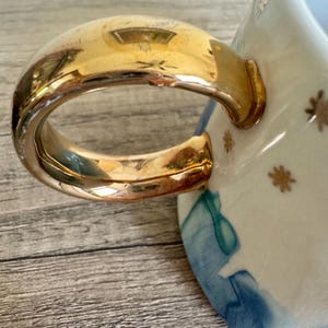 May include: A ceramic mug featuring a large, reflective gold-toned handle. The exterior of the mug is decorated with a pattern of small gold star icons against a white background, accented by abstract blue watercolor-style washes near the base.