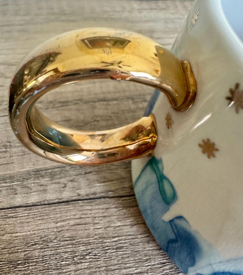 May include: A ceramic mug featuring a large, reflective gold-toned handle. The exterior of the mug is decorated with a pattern of small gold star icons against a white background, accented by abstract blue watercolor-style washes near the base.