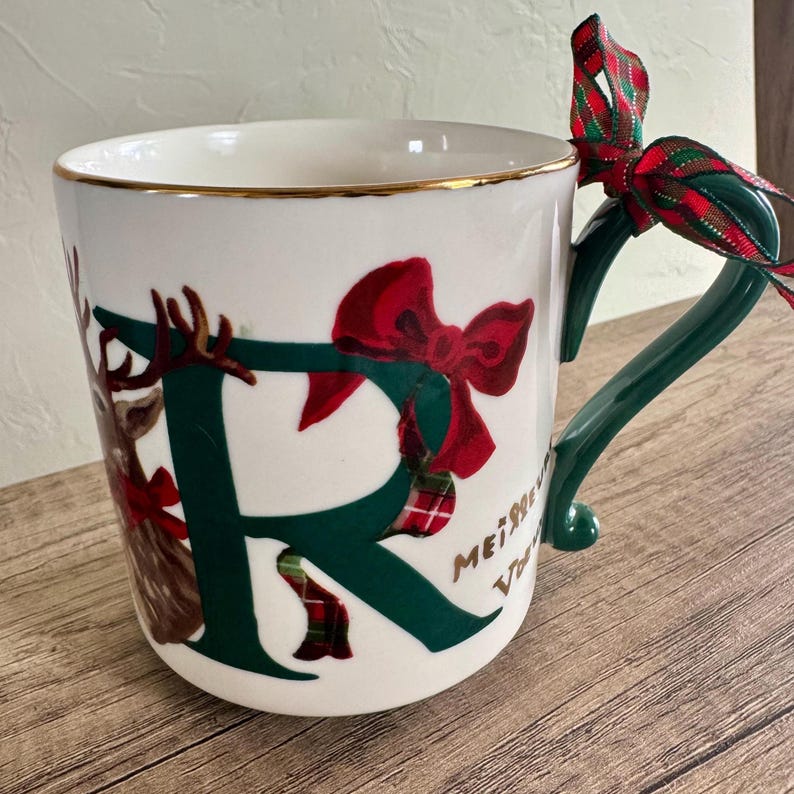 Puede incluir: Taza de cer&aacute;mica blanca con borde dorado, con una gran letra "R" verde decorada con un lazo rojo y una ilustraci&oacute;n de reno. El asa es verde y una cinta roja y verde est&aacute; atada al asa. La taza tiene las palabras "Merry" y "Vibes".