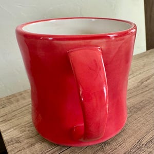 May include: A glossy red ceramic mug with a curved handle. The interior of the mug is white. The mug is sitting on a wooden surface.