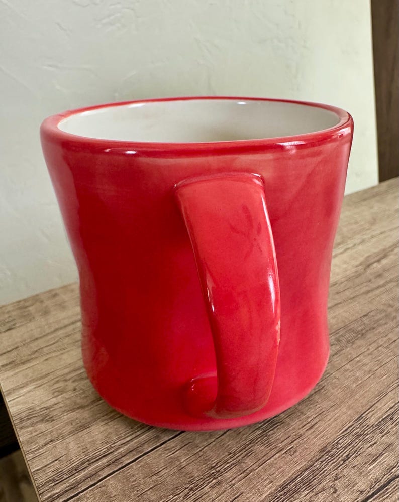 May include: A glossy red ceramic mug with a curved handle. The interior of the mug is white. The mug is sitting on a wooden surface.