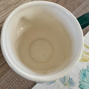 K&ouml;nnte beinhalten: Cremefarbene Keramik-Tasse mit einem dunkelgr&uuml;nen Henkel. Die Tasse hat eine weite &Ouml;ffnung und einen abgerundeten Boden. Der Innenraum ist heller cremefarben. Die Tasse steht auf einem Stoff mit Blumenmuster.