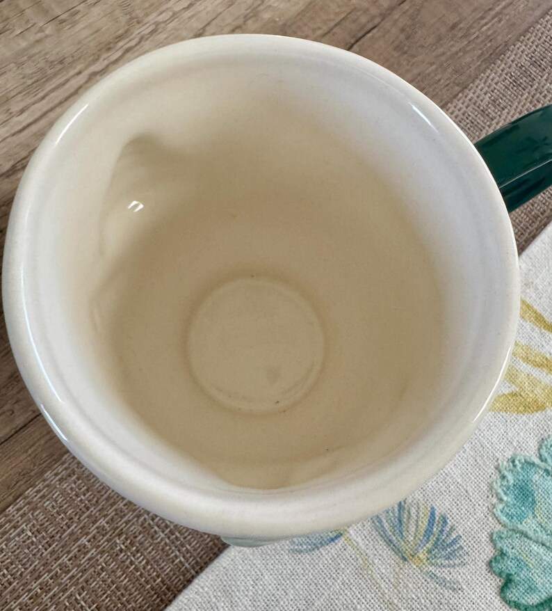 K&ouml;nnte beinhalten: Cremefarbene Keramik-Tasse mit einem dunkelgr&uuml;nen Henkel. Die Tasse hat eine weite &Ouml;ffnung und einen abgerundeten Boden. Der Innenraum ist heller cremefarben. Die Tasse steht auf einem Stoff mit Blumenmuster.