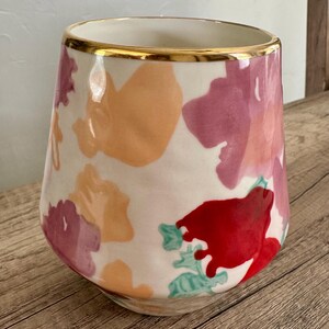 May include: A ceramic vase with a white base, decorated with abstract floral patterns in red, pink, and orange. The rim is trimmed with a gold band. The vase has a tapered shape and is suitable for home decor.