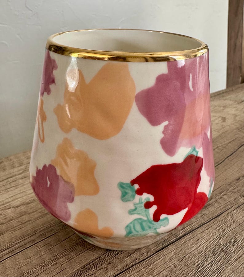May include: A ceramic vase with a white base, decorated with abstract floral patterns in red, pink, and orange. The rim is trimmed with a gold band. The vase has a tapered shape and is suitable for home decor.
