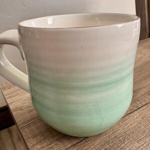 Pode incluir: Uma caneca de cer&acirc;mica grande com um design aquarela branco e verde. A caneca tem uma forma arredondada com uma al&ccedil;a e uma borda dourada. A cor verde se funde no branco, criando um efeito gradiente.