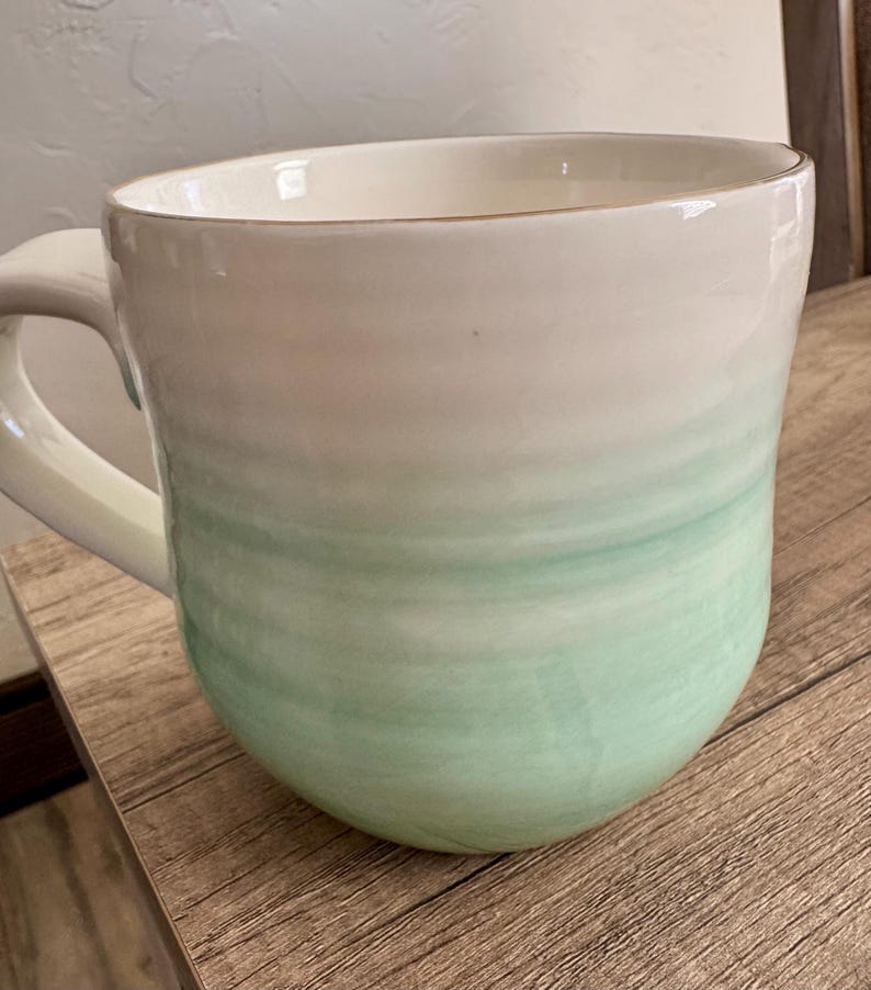 Pode incluir: Uma caneca de cer&acirc;mica grande com um design aquarela branco e verde. A caneca tem uma forma arredondada com uma al&ccedil;a e uma borda dourada. A cor verde se funde no branco, criando um efeito gradiente.