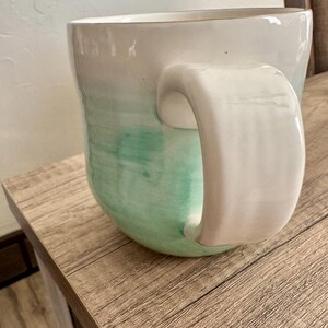 Pode incluir: Uma caneca de cer&acirc;mica com uma al&ccedil;a grande e curva. A caneca transita de branco no topo para um verde suave na base. A caneca est&aacute; sobre uma superf&iacute;cie de madeira.