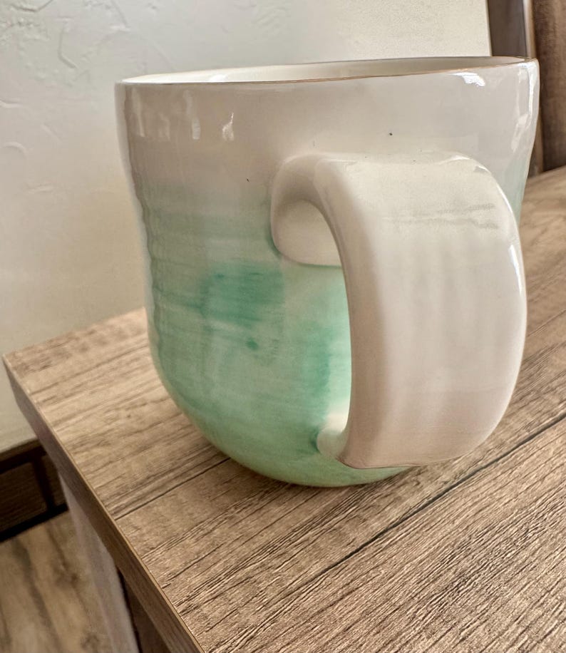 Pode incluir: Uma caneca de cer&acirc;mica com uma al&ccedil;a grande e curva. A caneca transita de branco no topo para um verde suave na base. A caneca est&aacute; sobre uma superf&iacute;cie de madeira.
