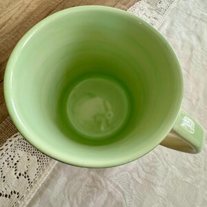 May include: A light green ceramic mug with a rounded interior and a handle. The mug's interior is a darker shade of green, and the handle has a white design. The mug is sitting on a white lace tablecloth.