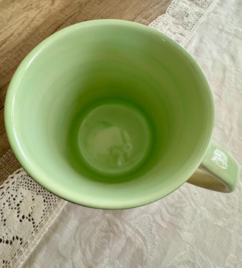 May include: A light green ceramic mug with a rounded interior and a handle. The mug's interior is a darker shade of green, and the handle has a white design. The mug is sitting on a white lace tablecloth.