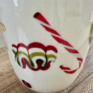 May include: White ceramic mug with a Christmas theme. The mug features a red and white candy cane design and a swirl of red, green, and white decorative elements. The mug is on a wooden surface.