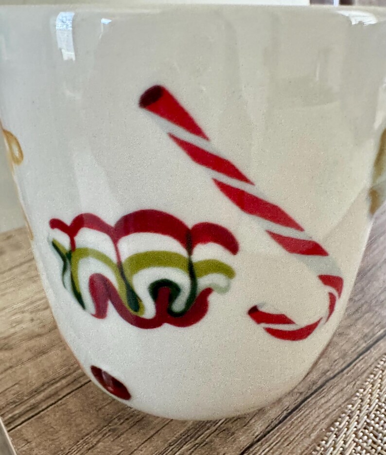 May include: White ceramic mug with a Christmas theme. The mug features a red and white candy cane design and a swirl of red, green, and white decorative elements. The mug is on a wooden surface.