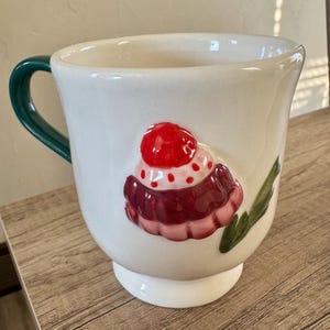 K&ouml;nnte beinhalten: Wei&szlig;e Keramik-Tasse mit gr&uuml;nem Henkel und Cupcake-Design. Der Cupcake hat eine rote Kirsche obenauf, mit rosa und kastanienbraunem Zuckerguss. Die Tasse steht auf einem wei&szlig;en Sockel.