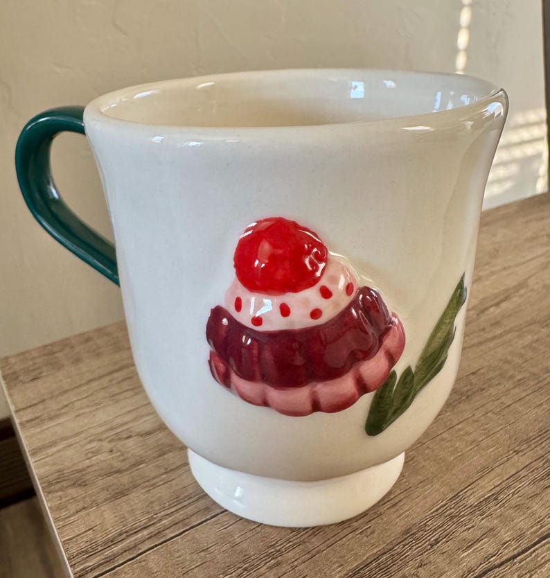 K&ouml;nnte beinhalten: Wei&szlig;e Keramik-Tasse mit gr&uuml;nem Henkel und Cupcake-Design. Der Cupcake hat eine rote Kirsche obenauf, mit rosa und kastanienbraunem Zuckerguss. Die Tasse steht auf einem wei&szlig;en Sockel.
