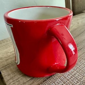 May include: A glossy red ceramic mug with a white interior and a large, curved handle. The mug has a white and black design on the side. The mug is sitting on a textured surface.