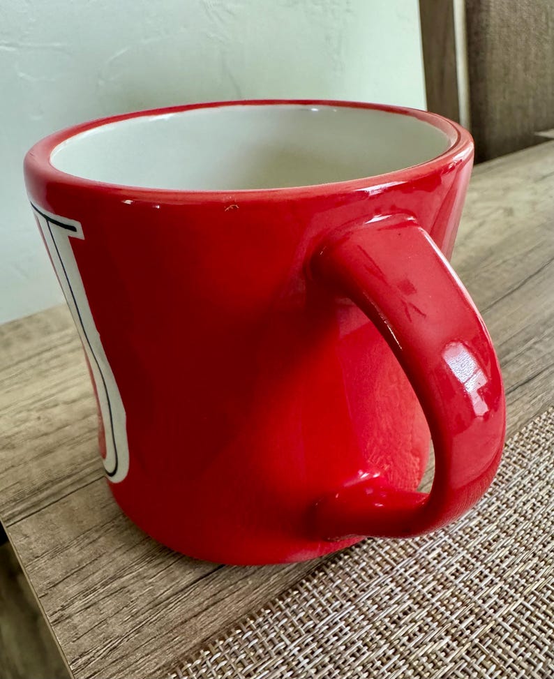May include: A glossy red ceramic mug with a white interior and a large, curved handle. The mug has a white and black design on the side. The mug is sitting on a textured surface.