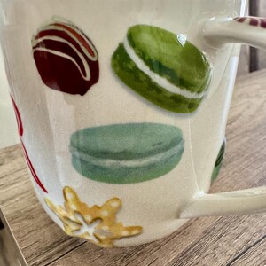 May include: Cream ceramic mug decorated with illustrations of a chocolate truffle, green and blue macarons, and a gold snowflake. The mug has a curved handle and is set on a wooden surface.